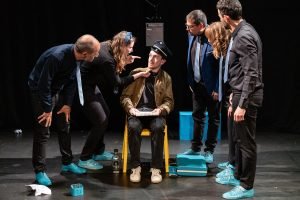 Une scène de cours de théâtre parisiens avec six acteurs interagissant sur scène, l'un assis et les autres debout autour de lui