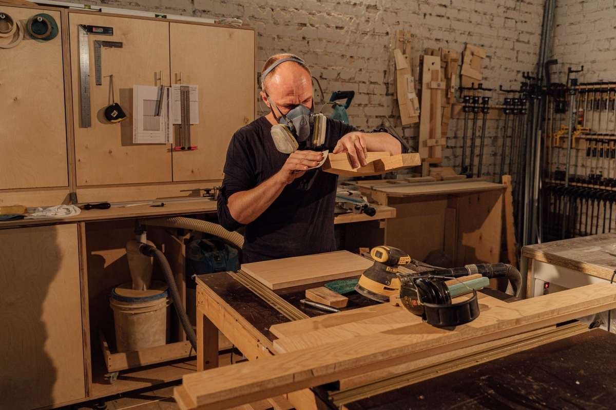 Homme portant masque, ponçant bois sur table dans un atelier rempli d'outils
